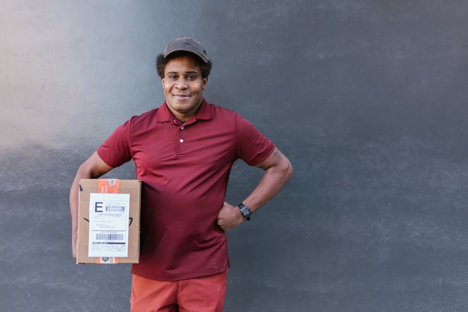 A smiling male delivery person with medium-dark skin, wearing a maroon polo shirt, red pants, and a dark cap, standing against a grey wall. He is holding a cardboard box with a shipping label in his l