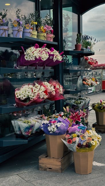 A flower display at a florist shop featuring an assortment of fresh floral arrangements in various colors and styles. On the upper shelf, small potted plants and pastel-colored bouquets wrapped in dec