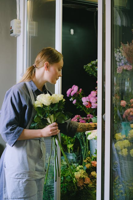 A young woman with light brown hair tied back, wearing a gray apron over a blue shirt, is selecting a bouquet of white roses at an indoor flower shop. She is holding the bouquet gently with both hands