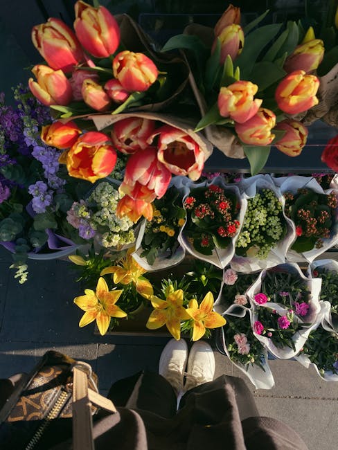 A top-down view of a variety of fresh flower bouquets arranged outdoors, displayed on a dark surface. The arrangements include a bouquet of peach and yellow tulips with green buds and long green leave