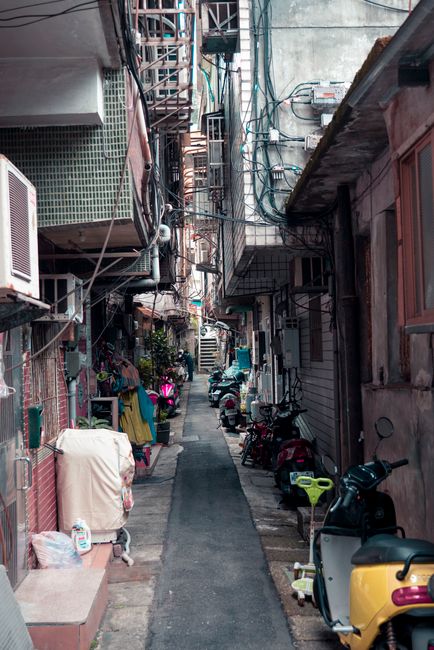 A narrow alleyway between tightly packed buildings featuring a paved pathway lined with various parked motorbikes, scooters, and bicycles. The buildings have a mix of metal and wooden balconies, some