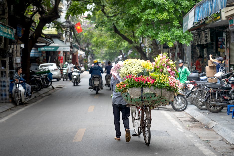 A street scene featuring a person pushing a bicycle laden with a large, vibrant floral arrangement. The bouquet includes various flowers such as pink and white lilies, yellow chrysanthemums, and clust