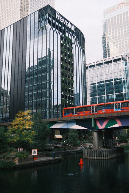 The image depicts a modern cityscape featuring a high-rise building with a reflective glass façade displaying the logo 'Revolut.' In front of the building, a vibrant orange double-decker train travel