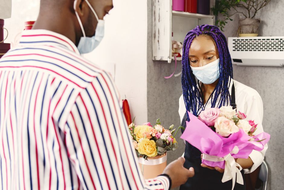 A professional florist at floristlimehouse.co.uk, with blue braided hair and wearing a face mask, is carefully arranging a bouquet of fresh pink roses, cream-colored roses, and small mixed flowers wra